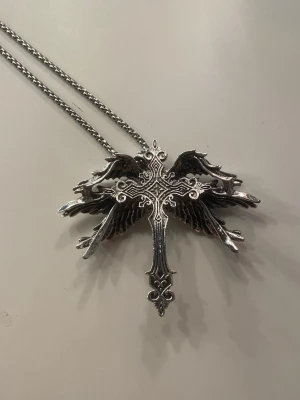 Silvrigt halsband med kors och vingar - Unikt halsband i silverfärgad metall med ett detaljerat kors och utbredda vingar. Kedjan är grov och smycket har en cool, gotisk vibe. Perfekt för dig som gillar statement-accessoarer och vill sticka ut med din stil. Helt oanvänd och relativt stor! ❤️