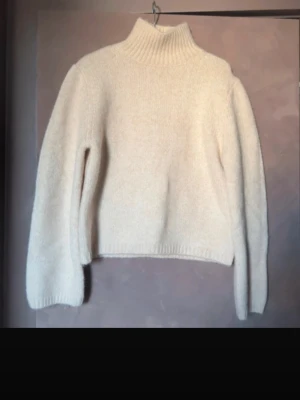 Beige stickad polotröja - Mysig beige stickad polotröja med lång ärm och ribbstickade muddar. Tröjan har en hög krage och är tillverkad i ett fluffigt, mjukt material som känns riktigt skönt mot huden. Perfekt för lager-på-lager och en clean look.
