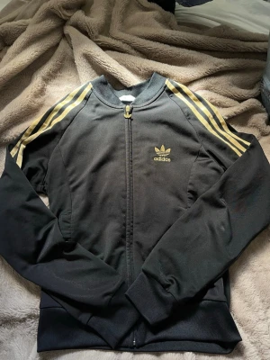 Svart Adidas track jacket med guld ränder - Svart track jacket från Adidas med klassiska guldiga ränder längs ärmarna och broderad logga på bröstet. Storlek 38. Obs ett litet hål på ärmen 