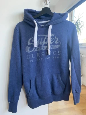 Blå hoodie från Superdry(M fits S) - Mysig blå hoodie från Superdry med trycket 'Superdry Classics' på bröstet. Har vit dragsko i huvan och en stor känguruficka framtill. Perfekt för en avslappnad och cool stil.
