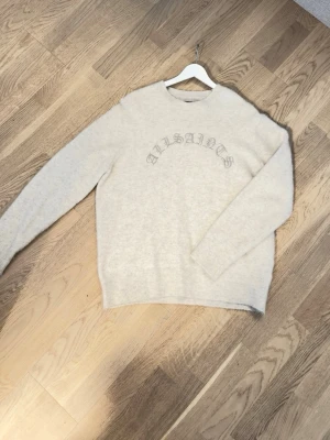 Grå/vit stickad tröja - Mysig grå/vit stickad tröja från AllSaints med rund halsringning och broderad logga i grått framtill.                      Köpt i London för ca 2k, använd typ 1 gång