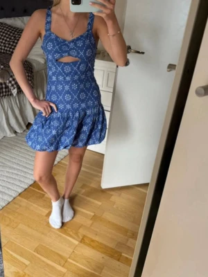  Ny  trendig Blå blommig kortklänning med cut-out - Söt blå kortklänning med vitt blommönster och volangkjol. Klänningen har breda axelband, en snygg cut-out detalj under bysten och markerad midja. Perfekt för varma dagar och somriga tillfällen.