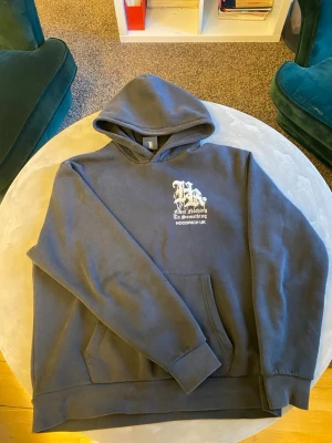 Svart hoodie från Hoodrich M - Svart hoodie från Hoodrich med vit tryckt logga och text på bröstet. Klassisk känguruficka framtill och stor huva. Mjuk insida och avslappnad passform, perfekt för en chill streetwear-look.