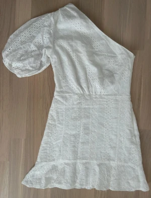 Vit oneshoulder klänning med volang - Superfin vit kortklänning i bomull med broderade detaljer och volangkant. Klänningen har oneshoulder-design och en puffig ärm, vilket ger en trendig och somrig look. Perfekt för varma dagar och speciella tillfällen. Jättefint skick🌸💕Perfekt till studenten!💗