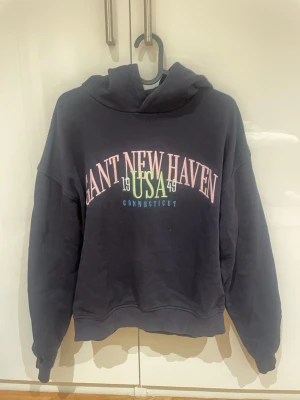 Mörkblå hoodie Gant New Haven - Mörkblå hoodie från Gant med tryck i rosa, grönt och vitt framtill där det står 'GANT NEW HAVEN USA 1949 CONNECTICUT'. Klassisk huva och ribbade muddar vid ärmslut och nederkant. Perfekt för en avslappnad och sportig stil.