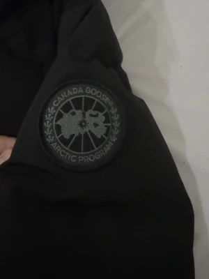 Svart dunjacka från Canada Goose - Säljer en svart dunjacka från Canada Goose med klassisk patch på ärmen. Jackan har huva, dragkedja och knappar framtill samt ribbade muddar vid ärmsluten. Perfekt för kalla vinterdagar och riktigt varm tack vare dunfyllningen.