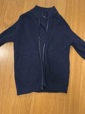 Mörkblå stickad kofta med dragkedja - Säljer en mörkblå stickad kofta med hel dragkedja och hög krage. Tröjan har ribbad struktur och är långärmad, perfekt för lager-på-lager. Enkel och stilren design som passar till jeans eller chinos.