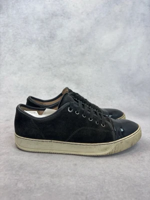 Svarta Lanvin sneakers med lackdetalj - Stilrena svarta sneakers med ovandel i mocka och glansig lackdetalj på tån. Skorna har svarta skosnören, metallöljetter och en vit sula med vintagekänsla. Perfekta för dig som gillar en clean look med en twist.