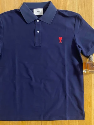 Mörkblå piké från Ami med rött hjärta - Snygg mörkblå pikétröja från Ami med klassisk krage och två knappar framtill. På bröstet sitter Ami:s ikoniska röda hjärtemblem. Tillverkad i mjuk bomull och har korta ärmar. Perfekt för en clean och stilren look.