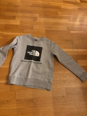 Grå sweatshirt från The North Face - Snygg grå sweatshirt från The North Face med klassisk logga och texten 'Never Stop Exploring' på bröstet. Tröjan har rund hals, långa ärmar och ribbade muddar vid ärmslut och nederkant. Perfekt för en avslappnad och sportig stil.