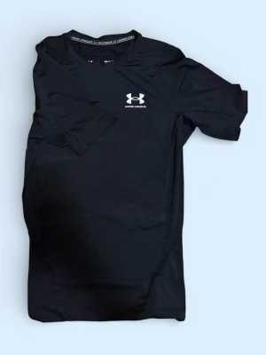 Svart träningströja Under Armour - Svart kortärmad träningströja från Under Armour med logga på bröstet och reflexdetalj i nacken. Tillverkad i ett stretchigt funktionsmaterial som andas, perfekt för intensiva träningspass eller löpning.
