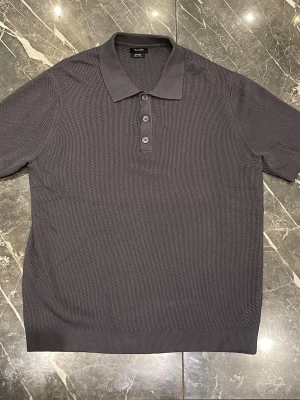 Mörkgrå pikétröja från Massimo Dutti - Stilren mörkgrå pikétröja från Massimo Dutti med klassisk krage och tre knappar framtill. Tröjan har en diskret strukturerad yta och är tillverkad i mjuk bomull, perfekt för en clean och avslappnad look. 