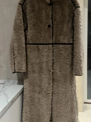 Beige teddyjacka från Gina Tricot - Lång, beige teddyjacka från Gina Tricot med svarta detaljer längs sömmarna. Jackan har en fluffig och mjuk yta, raka ärmar och knäppning framtill. Perfekt för kalla dagar när du vill vara både varm och snygg.