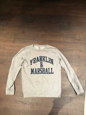 Grå sweatshirt från Franklin & Marshall - Klassisk grå sweatshirt med mörkblå Franklin & Marshall-tryck på bröstet. Tröjan har rund halsringning, långa ärmar och ribbade muddar vid ärmslut och nederkant. Perfekt för en avslappnad och sportig stil.