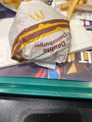 Double Cheeseburger från McDonald's - En klassisk Double Cheeseburger från McDonald's, inslagen i vitt papper med brunt och gult tryck. Perfekt för dig som gillar snabbmat och vill ha en rejäl burgare med dubbla lager ost och kött.