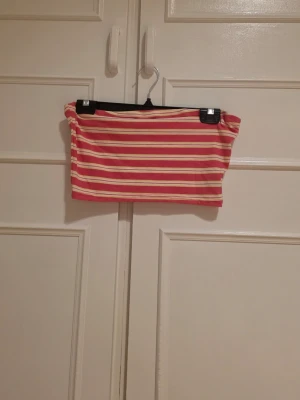 Randig rosa, gul och vit tubtopp - Säljer en randig tubtopp i rosa, gult och vitt. Toppen är ribbad och axelbandslös. Finns matchande kjol