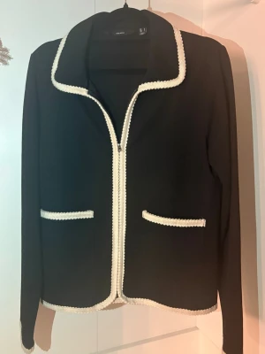 Svart kavaj med vita detaljer Vero Moda - Stilren svart kavaj från Vero Moda med vita virkade kanter längs krage, fickor och nederkant. Kavajen har två framfickor och klassisk krage, perfekt för en chic och trendig look. Materialet är mjukt och följsamt.