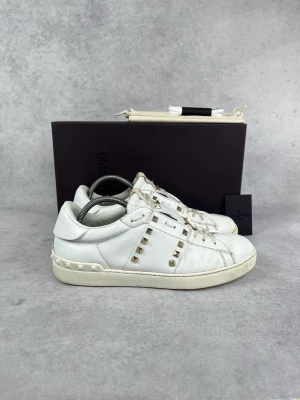 Vita Valentino sneakers med nitar - Snygga vita sneakers från Valentino med ikoniska guldfärgade nitar längs sidorna. Skorna har en klassisk låg siluett, rund tå och vita snören. Tillverkade i läder med detaljerad sula och diskret logga på plösen. Perfekta för dig som gillar exklusiv streetstyle.