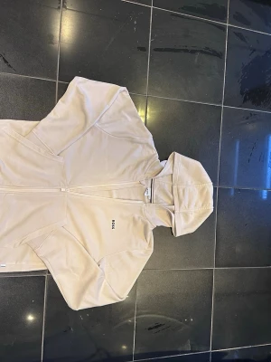 Beige hoodie från BOSS med dragkedja - Snygg beige hoodie från BOSS med dragkedja framtill och minimalistisk logga på bröstet. Hoodien har huva och är tillverkad i mjukt material som känns skönt mot huden. Perfekt för en avslappnad och stilren look.