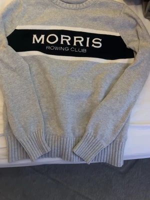 Grå stickad tröja från Morris - Snygg grå stickad tröja från Morris Rowing Club med bred svart och vit rand över bröstet och stor logga. Tröjan har rund halsringning och ribbade muddar vid ärmslut och nederkant. Perfekt för en stilren och sportig look.