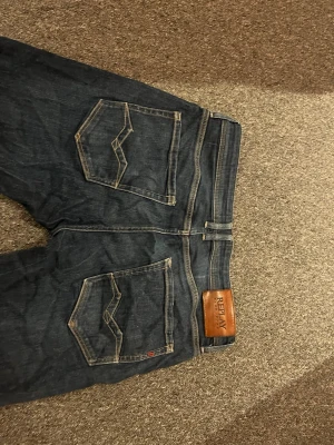 Mörkblå jeans från Replay - Säljer ett par mörkblå jeans från Replay med raka ben och klassiska kontrastsömmar. Jeansen har två bakfickor med unik söm och Replay-läderpatch i midjan. Tillverkade i slitstark denim, perfekta för en avslappnad och snygg stil.