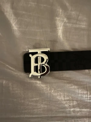 Svart Burberry bälte med TB-spänne - Snyggt svart bälte från Burberry med klassiskt rutmönster och ett stort, silverfärgat TB-spänne framtill. Bältet är gjort i skinn och har en stilren, modern look som passar till många outfits. SKRIV  OM NI HAR NÅGRA FUNDERINGAR 