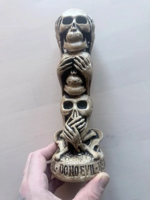 Dekorativ statyett med dödskallar - Cool prydnadsfigur i beige och svart föreställande tre dödskallar staplade på varandra, inspirerad av 'see no evil, hear no evil, speak no evil'. Tillverkad i resin med detaljerade ben och händer som täcker ögon, öron och mun. Perfekt för dig som gillar alternativ inredning. Prislapp på undersidan 480kr