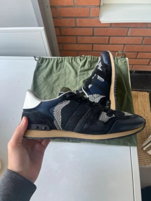 Valentino Garavani Rockrunner sneakers - Snygga Valentino Garavani Rockrunner sneakers i svart, blått och grått med vita detaljer. Ovandelen är i mesh och mocka med läderpaneler och ikoniska nitar vid hälen. Klassisk låg modell med beige sula och svarta skosnören. Perfekta för en trendig streetwear-look.