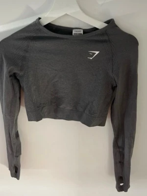 Mörkgrå cropped tröja från Gymshark - Snygg mörkgrå cropped långärmad tröja från Gymshark med ventilerande meshdetaljer och tumhål i ärmsluten. Tillverkad i stretchigt funktionsmaterial som passar perfekt till träning. Diskret vitt Gymshark-logga på bröstet.
