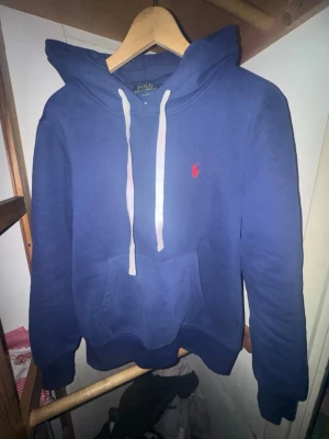 Blå hoodie från Polo Ralph Lauren - Snygg blå hoodie från Polo Ralph Lauren med vit dragsko och klassisk röd logga broderad på bröstet. Hoodien har en stor känguruficka framtill och ribbade muddar vid ärmslut och nederkant. Perfekt för en chill och stilren look.