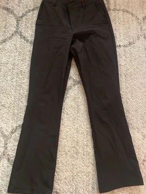 Svarta kostymbyxor med utsvängda ben - Stilrena svarta kostymbyxor med klassisk passform och utsvängda ben och låg midja. Byxorna har hällor för bälte och diskreta fickor framtill. Perfekta för en dressad look och enkla att matcha med olika toppar. 