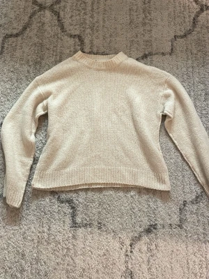Beige stickad tröja med rund hals - En enkel och stilren beige stickad tröja med rund halsringning och långa ärmar. Tröjan har ribbade muddar vid ärmslut och nederkant. Perfekt för lager på lager och passar till det mesta. Materialet ser ut att vara mjukt och bekvämt.