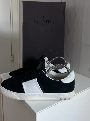 Valentino flycrew - Snygga sneakers från Valentino i svart mocka och vitt läder. Klassisk låg modell med vit sula och diskreta nitar på hälen. Skorna har svart ovandel med vita detaljer och svart plös. Perfekta för dig som gillar stilrena och exklusiva sneakers. 8 nitar borta sammanlagt, har lite hål bak och vid sidan som man kan se på bilderna, box två dusbag får man med pris kan diskuteras.