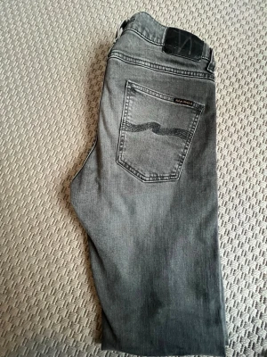 Grå jeans från Nudie Jeans - Snygga grå jeans från Nudie Jeans med klassisk femficksmodell och raka ben. Jeansen har en diskret logga på bakfickan och är tillverkade i mjuk denim. Perfekta för dig som gillar en stilren och avslappnad look. W30 L32
