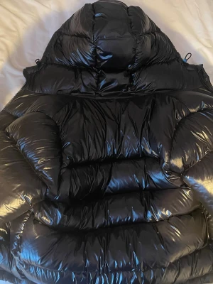 Svart glansig dunjacka med huva - Säljer en svart, glansig dunjacka med stor vadderad huva och breda kanaler. Jackan har en puffig siluett och dragsko i nederkant. Perfekt för kalla vinterdagar och riktigt varm tack vare tjockt dunfoder.