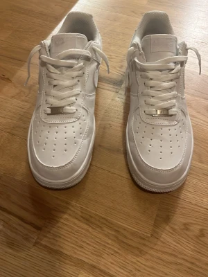 Nike Air Force 1 vita sneakers - Klassiska helvita Nike Air Force 1 sneakers i lågt utförande. Skorna har ovandel i läder, perforerade detaljer på tån och Nike-logga på sidan och hälen. Tjock vit sula och vita skosnören. Perfekta för en clean och stilren look.