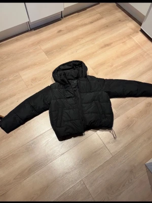 Svart pufferjacka med huva - Säljer en svart pufferjacka med huva och dragkedja framtill. Jackan har en boxig passform, två sidofickor och justerbar dragsko nedtill.  Nypris är 50€
