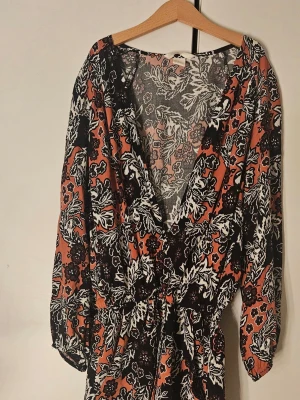 Blommig byxdress i orange och svart - Snygg byxdress med blommigt mönster i svart, vitt och orange. Byxdressen har markerad midja med knytband och djup v-ringning. Lätt och luftig i materialet, perfekt för dig som gillar boho-vibbar och färgstarka prints.