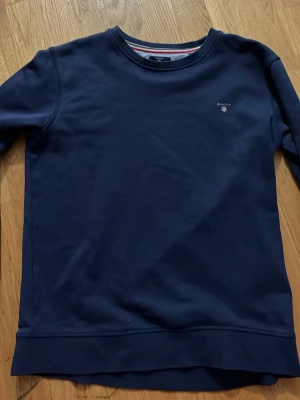 Mörkblå sweatshirt från GANT - Snygg mörkblå sweatshirt från GANT med rund halsringning och diskret logga broderad på bröstet. Tröjan har ribbade muddar vid ärmslut och nederkant. Perfekt för en clean och stilren look.