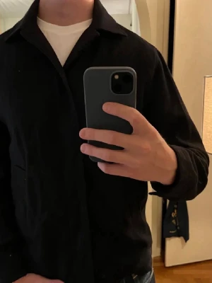 Marinblå overshirt från Peak Performance  - Stilren svart overshirt med klassisk krage och raka ärmar. Overshirten har en minimalistisk design utan synliga loggor eller detaljer, perfekt att bära över en t-shirt. Materialet ser ut att vara bomull eller en bomullsblandning. Pris kan diskuteras.