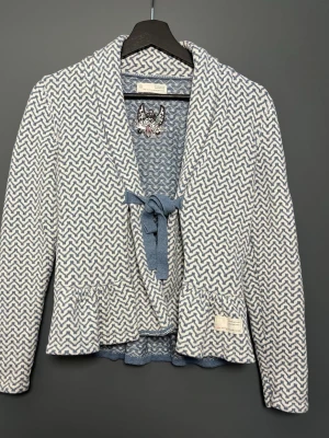 Mönstrad kofta från Odd Molly - Säljer en blåvit kofta från Odd Molly med zigzag-mönster och knytband framtill. Koftan har en avslappnad passform, volangkant nedtill och långa ärmar. Perfekt för dig som gillar unika detaljer och mjuka material.