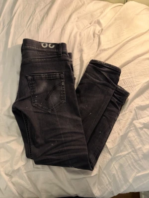 Dondup jeans - Säljer ett par svarta dondup jeans med slitningar och små färgstänk för en cool, avslappnad look. Jeansen har klassisk femficksmodell och smal passform. Perfekta för dig som gillar streetstyle och vill ha något unikt i garderoben.
