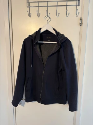 Massimo Dutti zip hoodie - En stilren mörkblå hoodie från Massimo Dutti med dragkedja framtill och två fickor. Hoodien har avtagbar huva med och är tillverkad i mjukt bomullsmaterial. Perfekt för en avslappnad look och enkel att matcha med andra plagg.