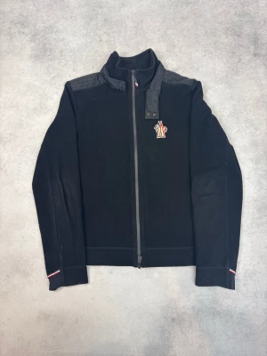 Svart fleecejacka från Moncler - Svart fleecejacka från Moncler med dragkedja framtill och hög krage. Jackan har en broderad logga på bröstet och quiltade detaljer på axlarna. Små rödvita detaljer vid ärmslut och krage ger extra stil.