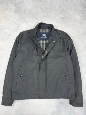 Svart jacka från Burberry med dragkedja - Stilren svart jacka från Burberry med klassiskt rutigt foder. Jackan har dragkedja och knappar framtill, två fickor med knappstängning och diskret krage. Perfekt för dig som gillar tidlös design och exklusiva detaljer.