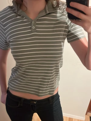 Gråvit randig polotopp - Kortärmad grå polotopp med vita horisontella ränder och knappdetaljer vid halsen. Toppen har en klassisk krage och är croppad i modellen, vilket ger en avslappnad och trendig look. Perfekt att matcha med jeans eller kjol.