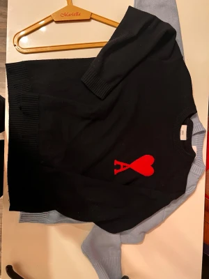 Svart stickad tröja från Ami Paris - Svart stickad tröja från Ami Paris med röd hjärtlogga och bokstaven A på bröstet. Tröjan har rund halsringning, ribbade muddar och är tillverkad i mjukt material. Perfekt för dig som gillar stilrena plagg med en twist.