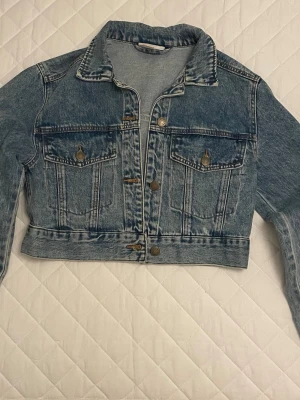 Cropped jeansjacka från Monki - Snygg croppad jeansjacka från Monki i tvättad blå denim. Klassisk modell med två bröstfickor, knappar framtill och krage. Perfekt för att lyfta vilken outfit som helst med en cool vintagekänsla.