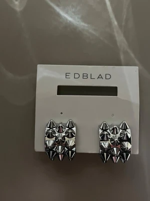 Silvriga nitar örhängen från Edblad - Säljer ett par coola silvriga örhängen från Edblad med fyrkantig form och dekorativa nitar i metall. Örhängena har en modern och edgy design som verkligen sticker ut. Perfekta för dig som vill ha något unikt och trendigt i smyckeskrinet. Aldrig använda! 