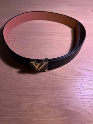 Louis Vuitton svart skärp med guldspänne - 105 cm, Snyggt svart skärp från Louis Vuitton med klassiskt monogrammönster och ett stort LV-spänne i guld. Bältet har en stilren design och insidan är beige. Perfekt accessoar för att lyfta vilken outfit som helst.